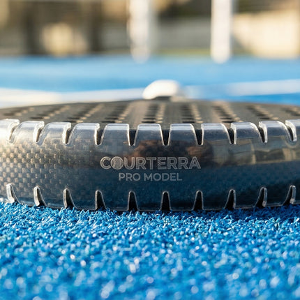 Premium padel racket with invisible Courterra Pro Model frame protection resting on a blue artificial grass padel court.
